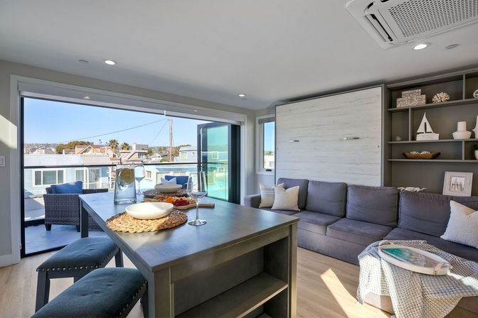 Bright beachside condo living room with a gray sectional, island dining table, sliding glass doors to a balcony and distant water and neighborhood views.