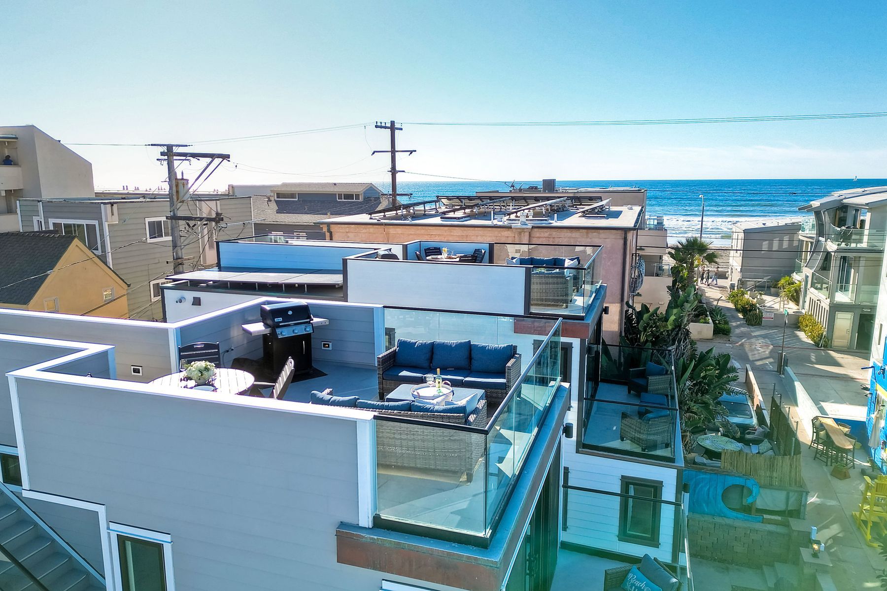 Sunny rooftop deck with glass railing, blue outdoor sofa and grill overlooking beach houses and the ocean in a coastal neighborhood.