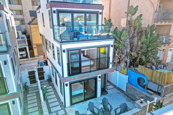 Three-story modern beach house with glass-railed balconies and rooftop lounge, sliding glass doors to a ground-level patio with Adirondack chairs, tropical palms and a narrow paved alley between neighboring homes.