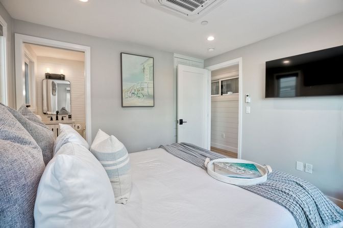 Coastal-chic master bedroom with white bedding, layered gray pillows and knit throw, decorative tray on the bed, wall-mounted TV, beach-themed artwork, and open door revealing a bright en-suite bathroom vanity.
