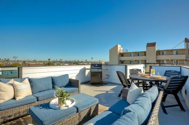 Sunny rooftop patio with blue-cushioned wicker sectional and ottoman, plant tray, gas grill and round dining table with chairs, overlooking an urban skyline with palm trees under a clear blue sky.