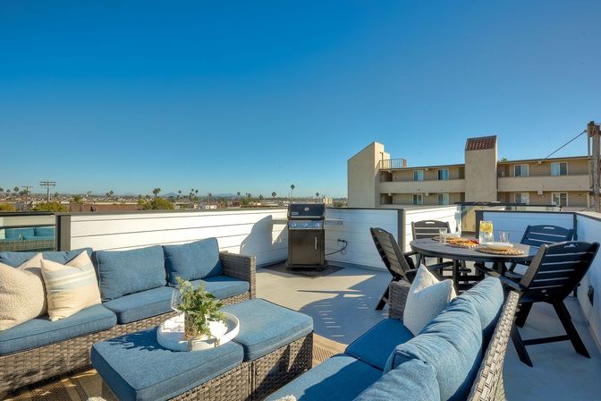 Sunny rooftop patio with blue-cushioned wicker sectional and ottoman, plant tray, gas grill and round dining table with chairs, overlooking an urban skyline with palm trees under a clear blue sky.