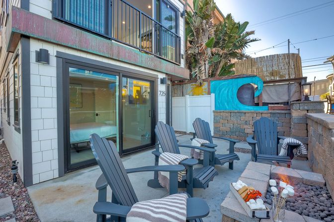 Cozy coastal patio at a vacation rental with Adirondack chairs and striped throws arranged around a stone fire pit stocked for s'mores, sliding glass doors to a bedroom, a colorful wave mural and tropical plants.