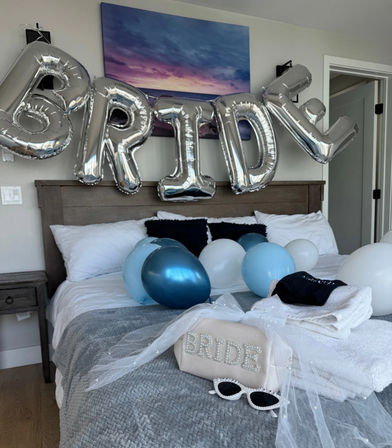 Silver foil balloons spelling BRIDE above a bed styled for a bride with blue and white balloons, a veil-draped pearl-embellished 'BRIDE' pouch, white towels and sunglasses on a gray quilt