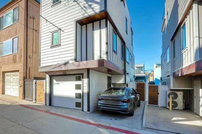 Contemporary white three-story townhouses lining a narrow urban alley, recessed garage with a gray sedan parked in the driveway, wood privacy gates and outdoor AC unit under a bright blue sky.