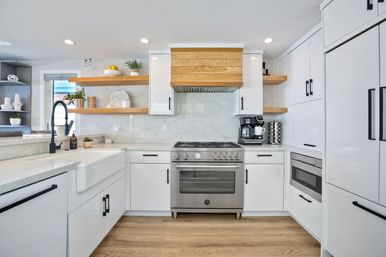 Bright modern white kitchen with marble backsplash, stainless steel gas range, farmhouse sink, black matte faucet, wood floating shelves and light oak floors.