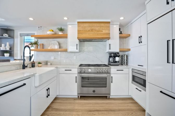 Bright modern white kitchen with marble backsplash, stainless steel gas range, farmhouse sink, black matte faucet, wood floating shelves and light oak floors.
