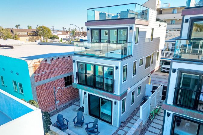 Modern three-story coastal townhouse with glass-railed balconies and rooftop deck, sliding glass doors to a ground-level patio with Adirondack chairs, narrow side path with a green bicycle, adjacent brick and stucco buildings under a clear blue sky
