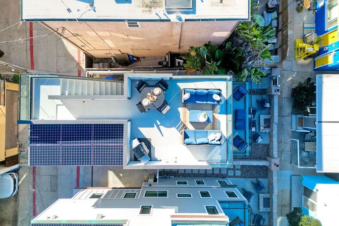 Bird's-eye view of a sunlit rooftop patio with solar panels, round dining table set for four, blue cushioned sofas, outdoor grill and tropical potted plants tucked between urban buildings.