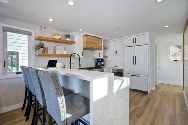 Bright modern kitchen with white marble waterfall island, three upholstered bar stools, floating wood shelves, black pull-down faucet, integrated white cabinetry and wood-look flooring.