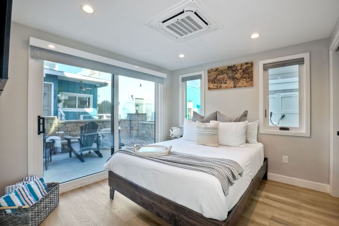 Bright coastal-style modern bedroom with white linens, layered pillows and gray throw, sliding glass doors opening to a sunny patio with Adirondack chairs and stone wall.