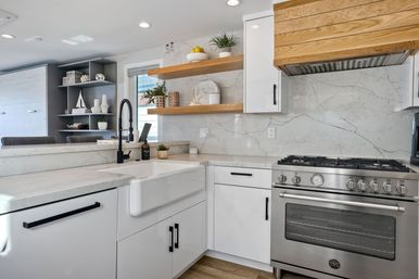 Bright modern open-concept kitchen with white farmhouse sink, matte-black pull-down faucet, marble backsplash, wood floating shelves and stainless-steel gas range