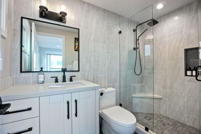 Sleek contemporary gray-tiled bathroom with glass walk-in shower and bench, white vanity with black matte faucets and large mirror
