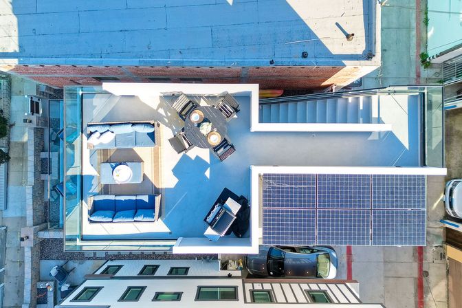 Aerial view of a modern urban rooftop patio — sunny seating area with blue cushions, round dining table, stairs, adjacent solar panels and a parked car below