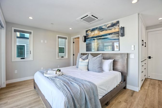 Modern coastal-inspired bedroom interior with a king bed, gray wood headboard, layered blue-gray pillows and knit throw, large ocean canvas above the bed, light hardwood floors, recessed lighting and built-in white cabinetry