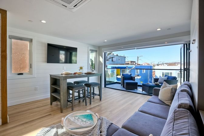 Sunny modern beachside living room with gray sectional, wall-mounted TV, high-top island and stools; folding glass doors open to a furnished balcony overlooking colorful seaside houses.