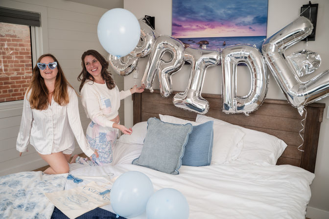Two women in pajamas celebrating a bride-to-be in a cozy coastal bedroom with silver "BRIDE" letter balloons and light blue balloons on a white bed.