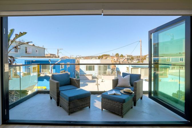 Sunny balcony with wicker lounge chairs and ottomans, hat and magazine on a tray, glass railing overlooking colorful houses and palm trees under a clear blue sky