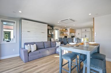 Sunny coastal-style open-concept living room and kitchen with a gray sofa before a closed white wood Murphy bed, light wood floors, a blue high dining table with stools set with glasses and a pitcher, and a bright modern kitchen island with bar stools.