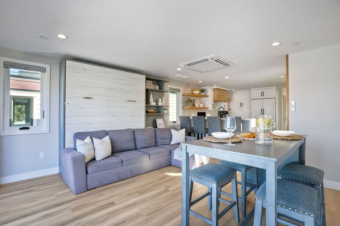 Sunny coastal-style open-concept living room and kitchen with a gray sofa before a closed white wood Murphy bed, light wood floors, a blue high dining table with stools set with glasses and a pitcher, and a bright modern kitchen island with bar stools.