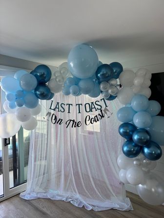 Coastal-themed balloon arch in shades of blue, teal and white with a "Last Toast On The Coast" banner hanging over a sparkly sequin backdrop beside sliding glass doors.