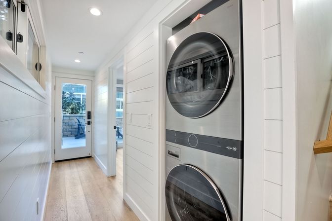 Bright narrow hallway in a modern home with a stacked stainless-steel washer and dryer in a recessed laundry nook, white shiplap walls, light hardwood floors, and a glass door leading to a small patio.