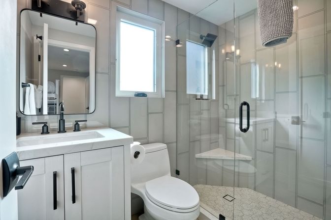 Bright modern bathroom with glass walk-in shower, white vanity and matte black fixtures, tiled walls, hexagon tile floor and a small window letting in natural light