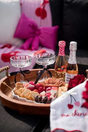 Romantic outdoor patio picnic tray with two glass coupe glasses tied with black ribbons, two mini rosé bottles, charcuterie platter of crackers, salami, berries and chocolates, a red rose and pink cushions in the background.