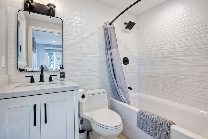 Bright modern white bathroom with textured wall tile, tub-shower combo with gray curtain, marble-top vanity, black fixtures and mirror.