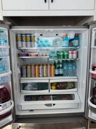 Open stainless-steel kitchen refrigerator filled with assorted beverages: canned seltzers and sparkling water on top, bottled water and sports drinks on the right, colorful canned drinks on a middle shelf, and a produce drawer with lemons and berries.