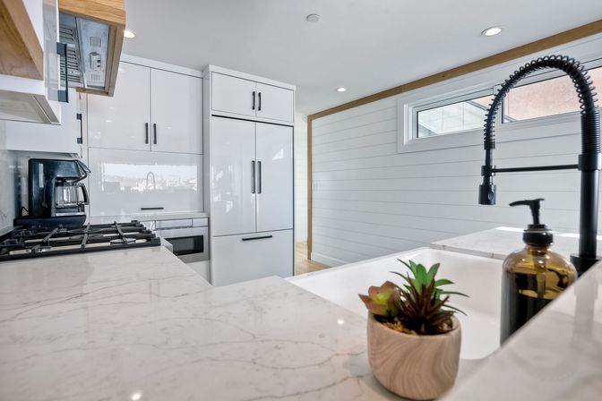 Bright, sleek modern white kitchen with marble countertops, integrated refrigerator, gas range, black spring pull-down faucet and a small potted succulent by the sink.