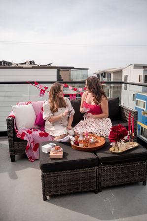 Two friends on an urban rooftop balcony sharing champagne amid Valentine's decor — wicker sofa with pink heart blanket, tray of bottles and desserts, red roses and candles.