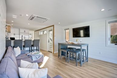 Sunlit coastal-style open-concept living room and kitchen with gray sectional sofa, white kitchen island with three bar stools, tall gray dining table with matching stools, wall-mounted TV, shiplap walls and light wood floors.