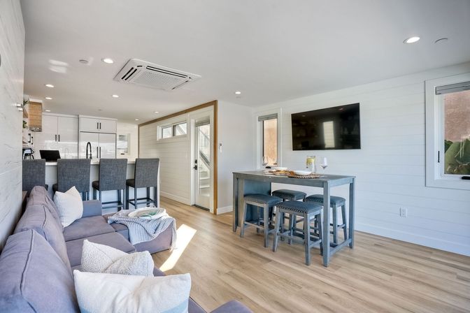 Sunlit coastal-style open-concept living room and kitchen with gray sectional sofa, white kitchen island with three bar stools, tall gray dining table with matching stools, wall-mounted TV, shiplap walls and light wood floors.