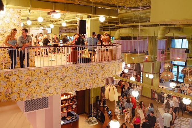 Two-story indoor bar and restaurant with a busy mezzanine balcony, floral wallpaper panels, hanging globe and wicker pendant lights, and crowds mingling on both floors