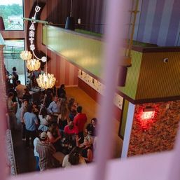 Mezzanine view of a bustling urban bar interior with groups mingling under chandeliers, a vertical neon 'ARTS' sign, and colorful striped walls