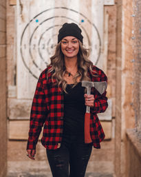 Smiling woman in a black beanie and red plaid shirt holding an axe at an indoor axe-throwing lane with a wooden bullseye target behind her.