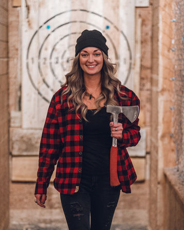 Smiling woman in a black beanie and red plaid shirt holding an axe at an indoor axe-throwing lane with a wooden bullseye target behind her.