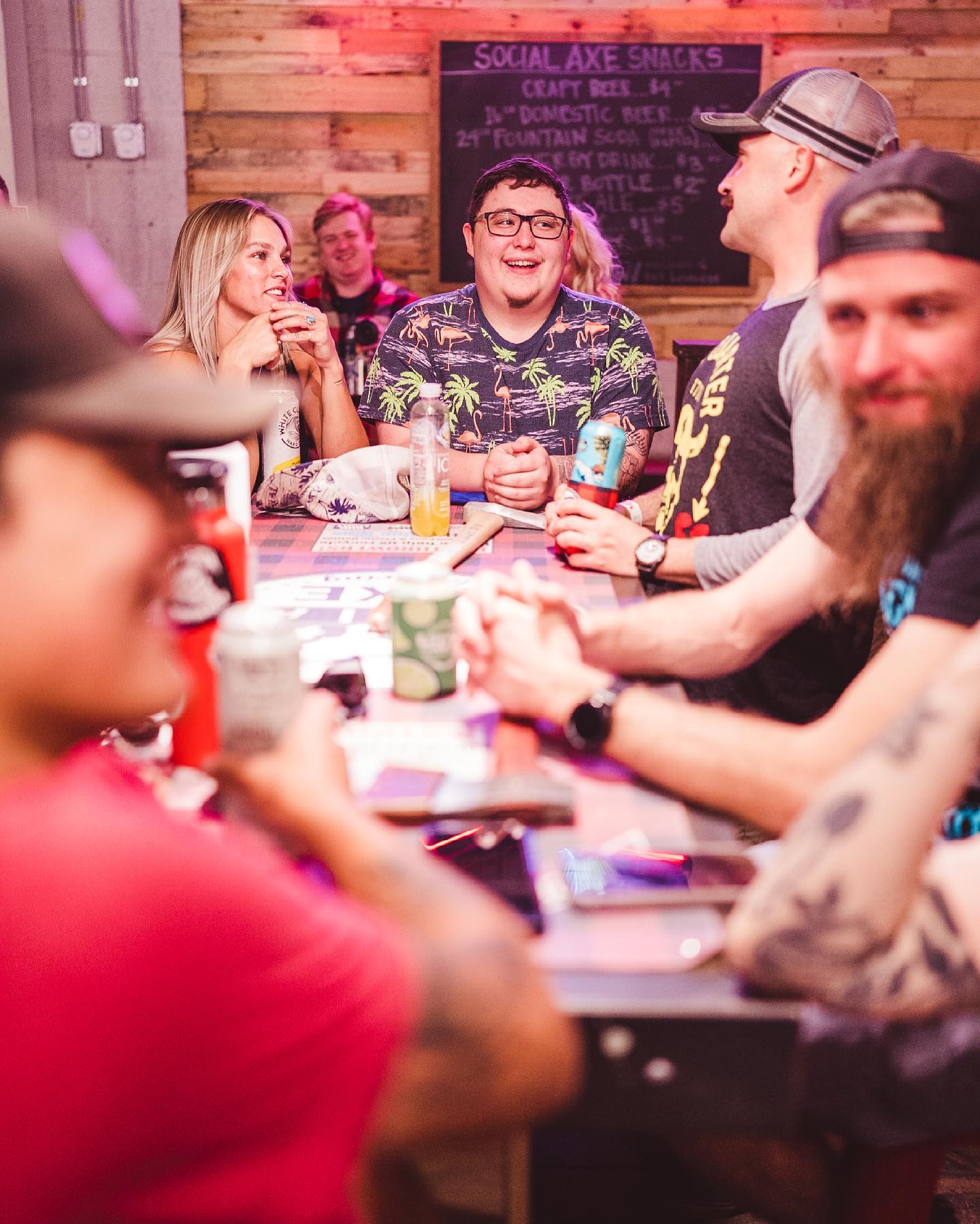 Group of friends laughing around a wooden high-top table at a rustic indoor bar with a chalkboard menu and colorful lighting, drinks and cans on the table — lively nightlife scene.