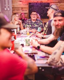 Group of friends laughing around a wooden high-top table at a rustic indoor bar with a chalkboard menu and colorful lighting, drinks and cans on the table — lively nightlife scene.