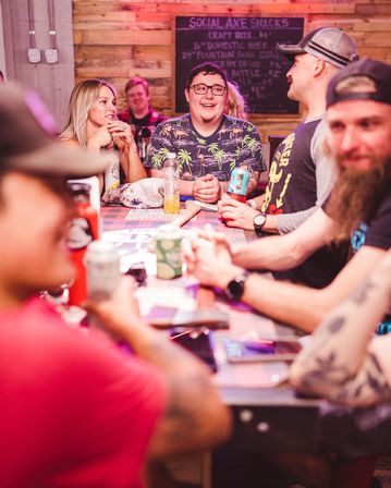 Group of friends laughing around a wooden high-top table at a rustic indoor bar with a chalkboard menu and colorful lighting, drinks and cans on the table — lively nightlife scene.