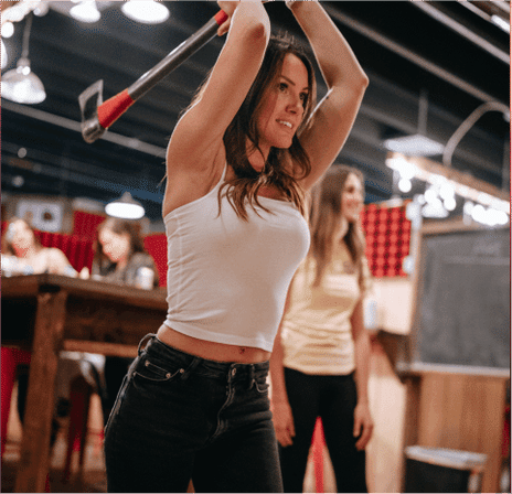 Woman mid-swing throwing an axe at an indoor axe-throwing lane, wearing a white tank top and dark jeans with friends watching — lively social outing