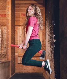 Person in a red shirt mid-jump holding a red-handled axe between their legs inside an indoor axe-throwing lane with wooden target boards in the background