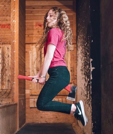 Person in a red shirt mid-jump holding a red-handled axe between their legs inside an indoor axe-throwing lane with wooden target boards in the background