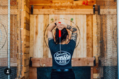 Rear view of a tattooed person raising a red-handled axe to throw at a wooden bullseye target inside an indoor axe-throwing lane with chain-link fencing.