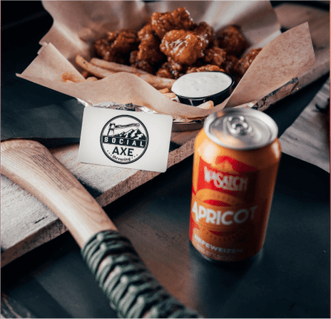 Rustic pub-style snack: basket of crispy chicken bites and fries with dipping sauce on parchment, apricot-flavored soda can and wooden axe handle on a serving board.