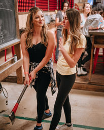 Two women at an indoor axe-throwing range, one holding an axe while they chat in a rustic wood-paneled recreation space with onlookers