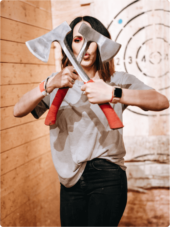 Person playfully holding two crossed red-handled axes in front of their face inside an indoor axe-throwing lane with wood-paneled walls and a bullseye target in the background.