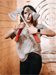 Person playfully holding two crossed red-handled axes in front of their face inside an indoor axe-throwing lane with wood-paneled walls and a bullseye target in the background.
