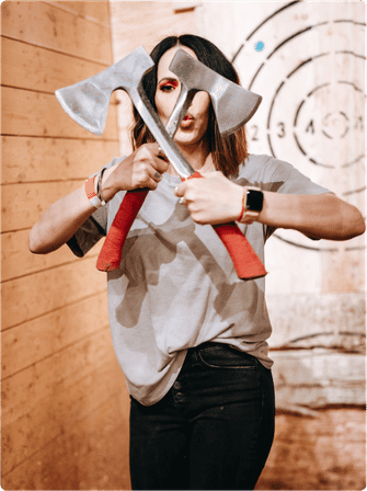 Person playfully holding two crossed red-handled axes in front of their face inside an indoor axe-throwing lane with wood-paneled walls and a bullseye target in the background.